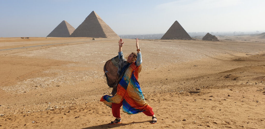 An expressive photograph of Sarah Hope standing in a dynamic, celebratory yoga posture on the desert sands of the Giza Plateau in Egypt. She wears vibrant, flowing orange, red, and blue fabrics that catch the desert breeze, with her arms raised joyfully toward the sky. In the background, the three iconic Great Pyramids of Giza rise majestically against a hazy, pale blue desert sky, showcasing the vast expanse of the ancient landscape.
