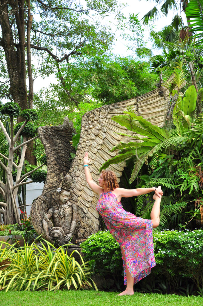 Yoga teacher Sarah Hope performing Natarajasana (Dancer Pose) in a lush tropical garden in Thailand with large stone wings in the background.
