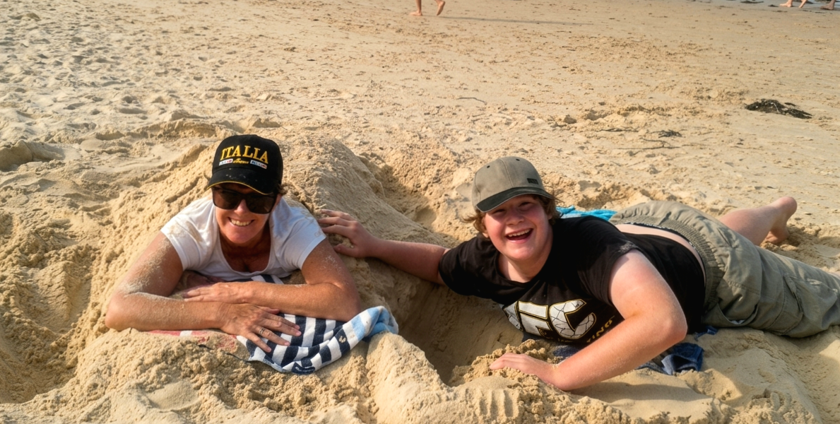 Sarah Hope and her son laughing while partially buried in the sand at the beach.