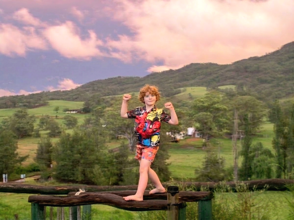 Young boy in a Spider-man shirt flexing his muscles while balancing on a rustic wooden bridge at sunset.