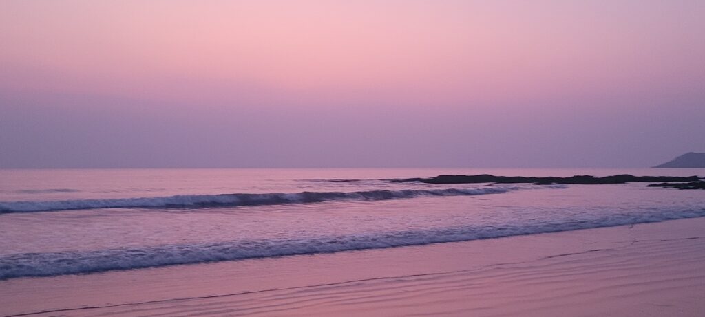 A peaceful, wide-angle view of gentle ocean waves washing onto a sandy beach under a soft violet and pink twilight sky, evoking the serene energy of the crown chakra.