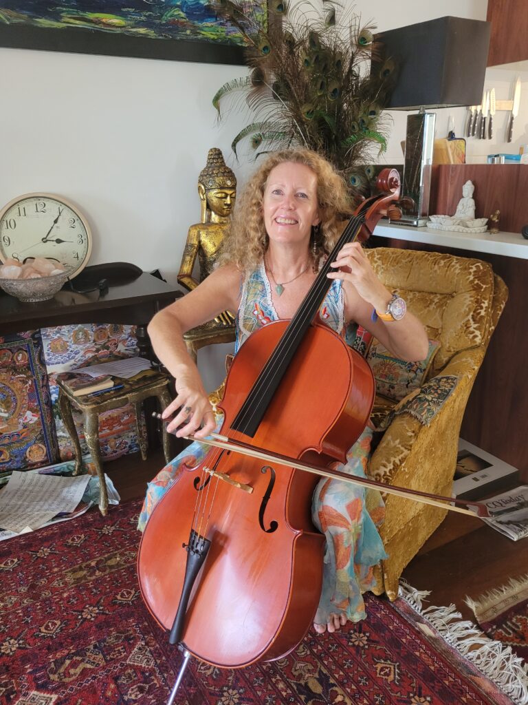 A warm, full-length photograph of Sarah Hope sitting comfortably in a gold velvet armchair, smiling warmly while playing a polished wooden cello. She is barefoot, wearing a colorful, flowing dress. Her home studio is filled with sacred art and decor, including a prominent golden Buddha statue behind her, an assortment of peacock feathers, a classic analog wall clock, a bowl of pink Himalayan salt crystals, and a detailed traditional thangka painting resting near a small side table on a patterned red Persian rug.