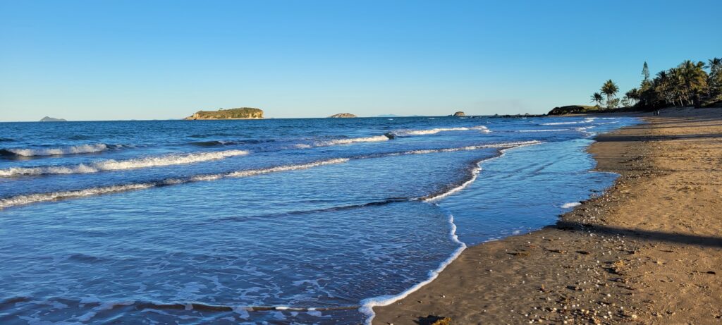 A wide landscape photograph of a serene coastal beach under a clear, bright blue afternoon sky. Gentle, low-cresting ocean waves roll softly onto a wide, sandy shoreline on the right, which is lined with tropical palm trees in the distance. Across the calm turquoise sea, a prominent flat-topped rocky island and smaller offshore islets rest quietly on the horizon under absolute, cloudless atmospheric clarity.