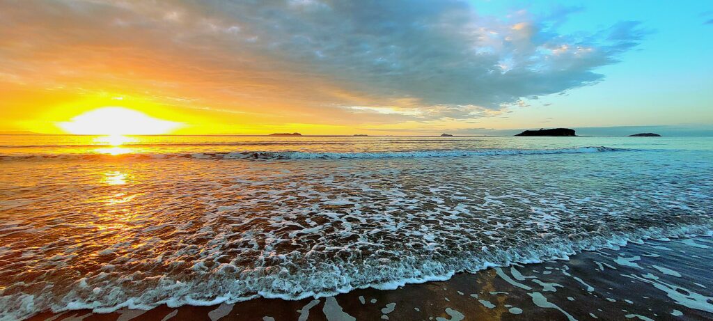 A wide-angle landscape photograph of a coastal sunrise over a calm ocean beach. On the left, a blazing golden-orange sun breaks through low clouds, casting a vibrant fiery reflection across the wet sand and rolling surf waves. On the right, the sky clears into a tranquil, soft blue above dark offshore islands on the horizon, with sea foam gently washing onto the shoreline in the foreground.