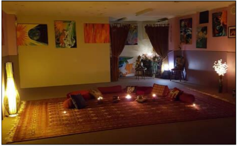 A cozy, dimly lit yoga and meditation studio space featuring a large patterned rug adorned with floor cushions, pillows, and glowing candles arranged for a group practice. Warm, ambient lamps illuminate the room, and colorful spiritual artwork hangs on the walls.